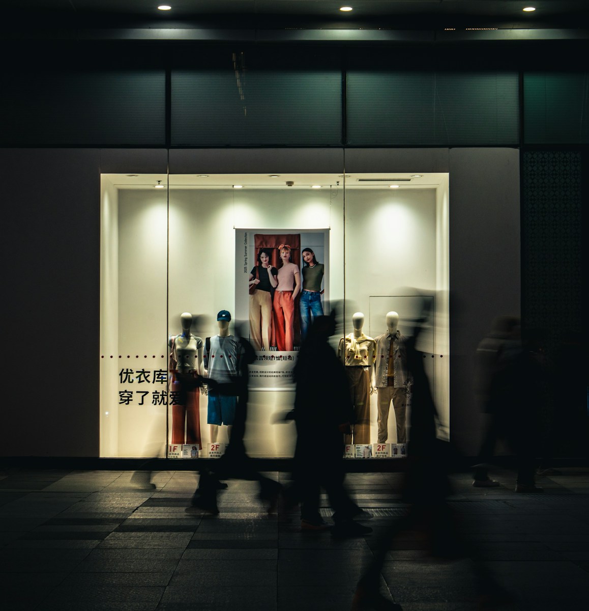 People walk past a brightly lit storefront at night.